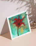 A mini art print of a red floral design on a turquoise background, displayed against a neutral backdrop with a shadow cast over it.
